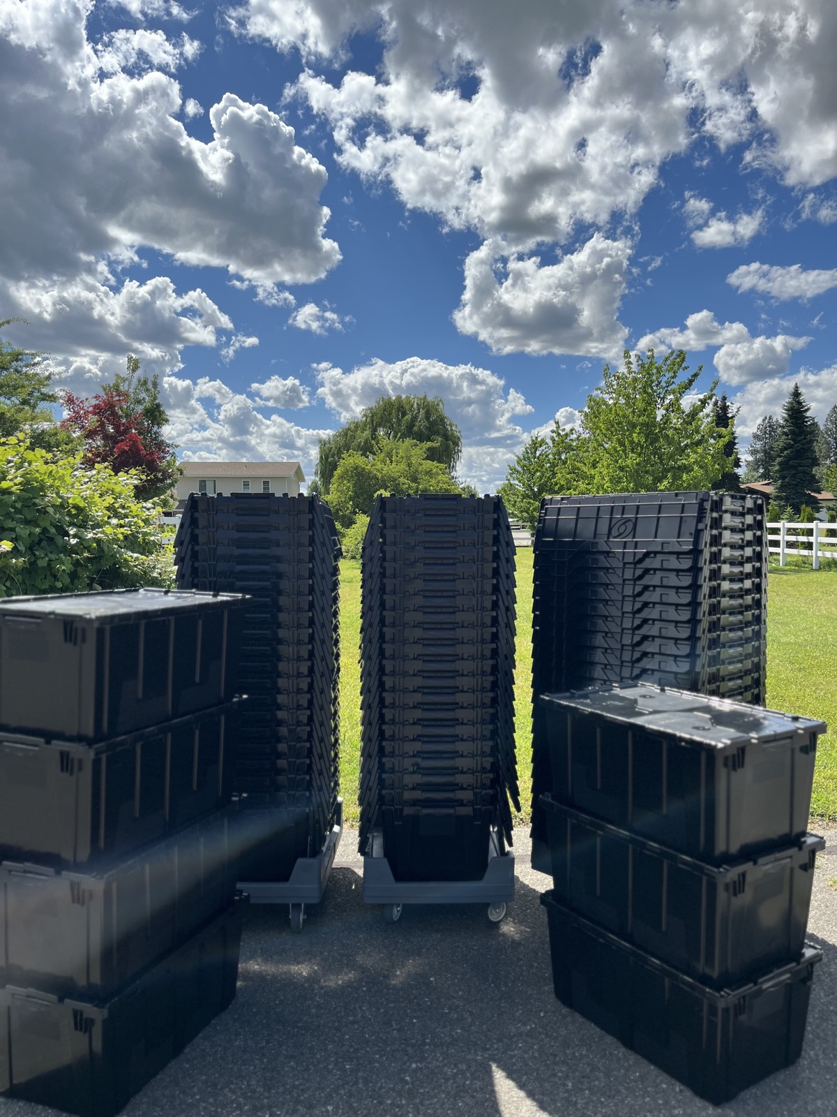 Rows of Tote Movers black totes stacked on a Coeur d'Alene driveway under a blue sky with cumulus clouds
